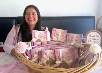 Smiling woman seated behind a wicker basket filled with pink gift boxes tied with cream ribbons. Advancing gifts are arranged on a table covered with a pink cloth.