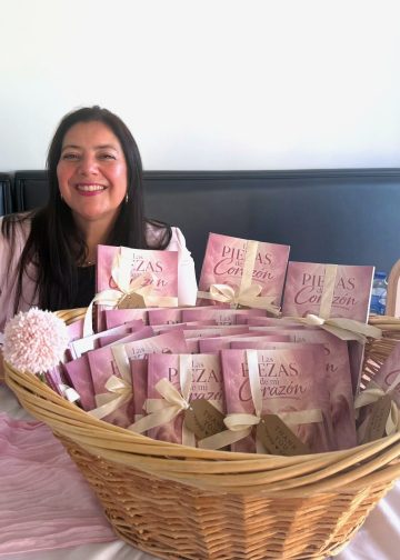 Smiling woman seated behind a wicker basket filled with pink gift boxes tied with cream ribbons. Advancing gifts are arranged on a table covered with a pink cloth.