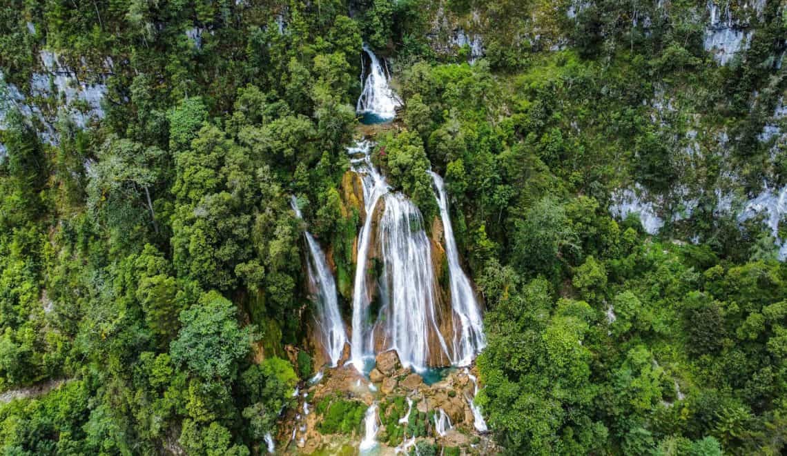 Aerial view of a multi-tier waterfall cascading down a rocky cliff through dense green forest into a turquoise pool below.