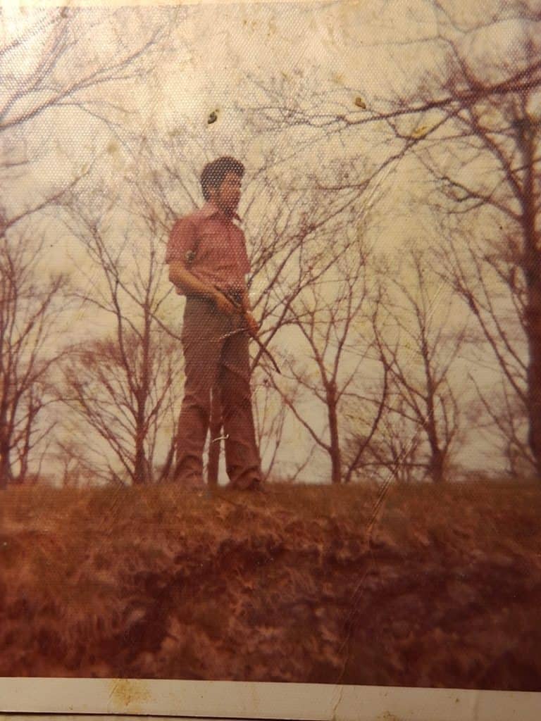 Man wearing brown clothes stands on a small hill in a leafless forest, holding a long-handled tool.