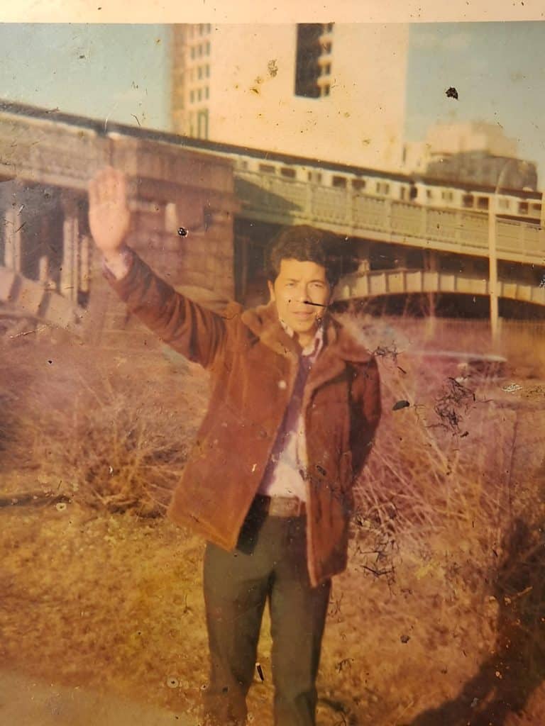 Man in a brown jacket waving with his right hand, outdoors near a concrete overpass or bridge.