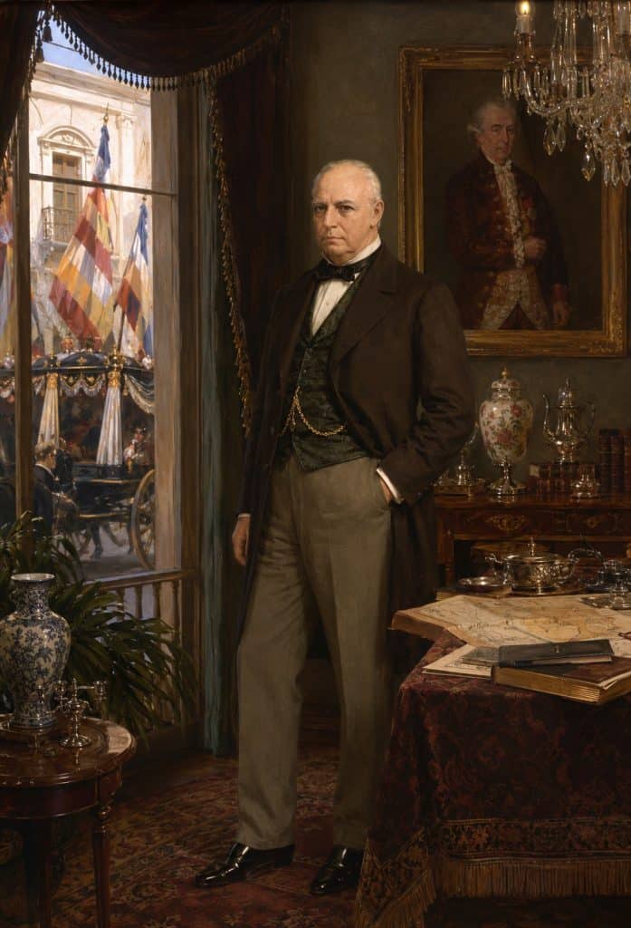 A distinguished older man in a dark coat and vest poses in a richly furnished 19th‑century room, with a chandelier, large portrait, and ornate tableware nearby.