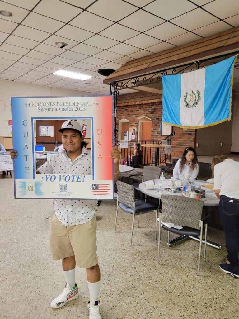 Man wearing a cap and white shirt holds a large photo-frame prop that says ¡Yo Voto! at a Guatemala-themed polling station, with a Guatemalan flag in the background.