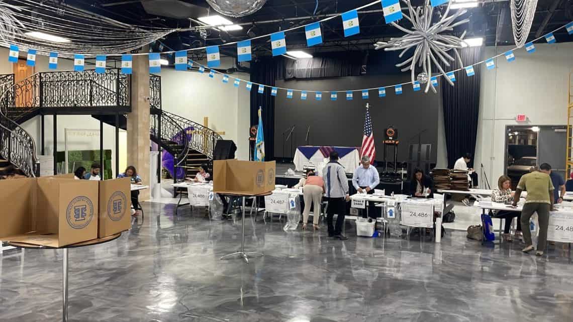 Indoor election polling site: volunteers sit at long tables processing ballots, with large cardboard ballot boxes and signs, a stage with flags in the background, and decorative blue-and-white banners overhead.