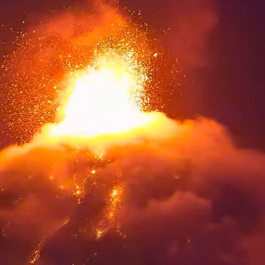 Volcanic eruption with a bright lava plume bursting above glowing clouds at night.