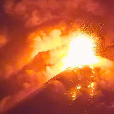Volcano eruption with a bright lava fountain and glowing orange clouds above a crater at nightfall/sunset.