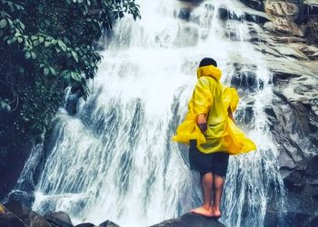 Person in a bright yellow raincoat stands barefoot on a rock, facing a multi-tiered waterfall in a lush, rocky setting.