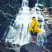 Person in a bright yellow raincoat stands barefoot on a rock, facing a multi-tiered waterfall in a lush, rocky setting.