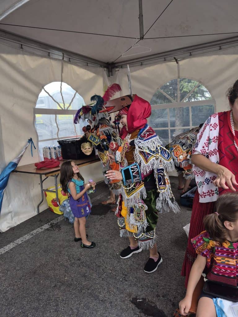 A child in a purple dress stands next to a performer wearing a bright, fringed ceremonial costume inside a white event tent.