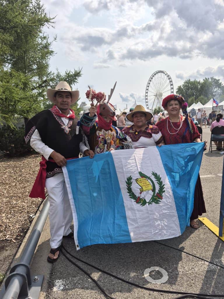 Four people in traditional clothing hold the blue‑and‑white Guatemala flag at an outdoor festival; a ferris wheel is visible in the background.