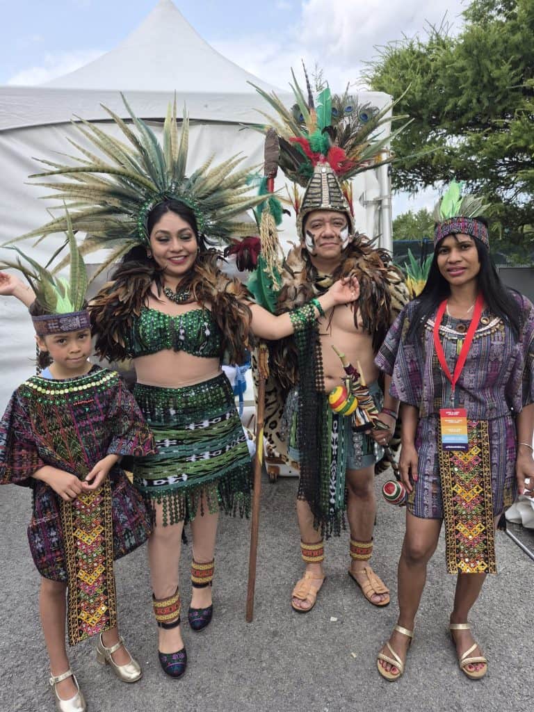 Group of four people in festive costumes with large feathered headdresses at an outdoor event, posing for a photo.