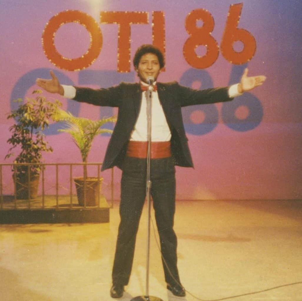 Male singer on a stage with arms outstretched, wearing a black suit and red cummerbund, microphone in front, pink/purple backdrop with large letters.”] ,