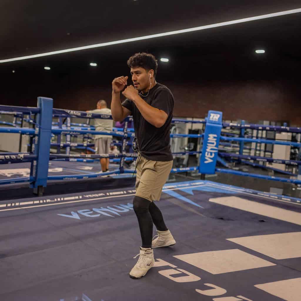 Male boxer in a ring, fists up in a guarding stance, wearing a black T-shirt, tan shorts, white sneakers, and wired earphones.