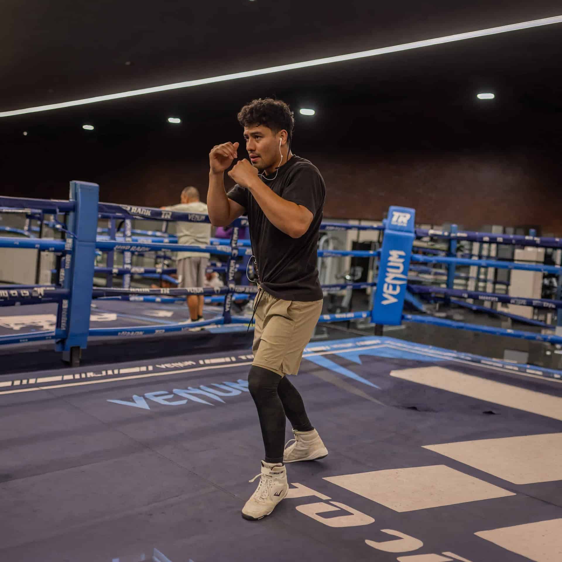Male boxer in a ring, fists up in a guarding stance, wearing a black T-shirt, tan shorts, white sneakers, and wired earphones.