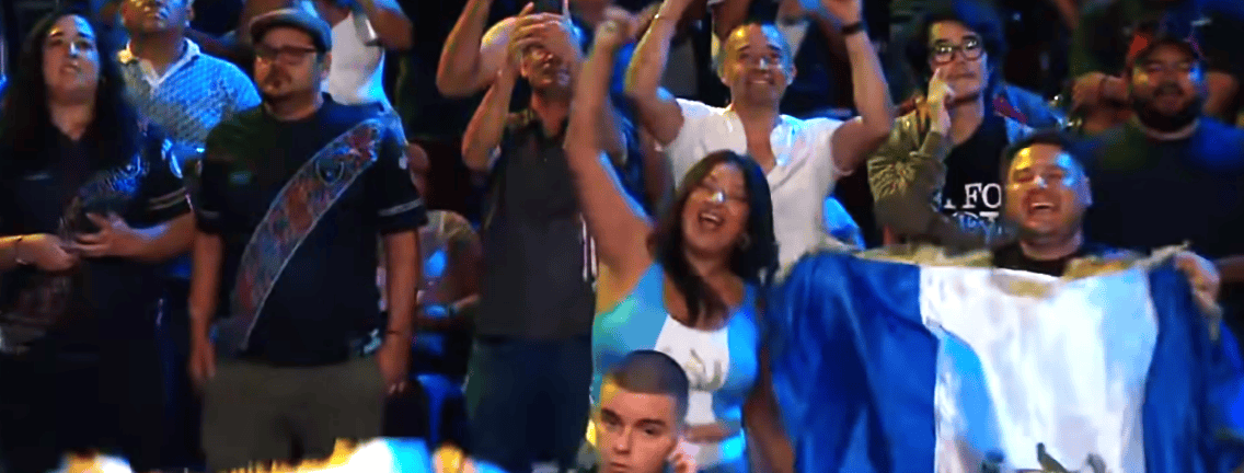 Crowd of cheering fans at a live event; a smiling woman in a blue top raises her arms while others cheer, with a blue-and-white flag visible nearby.