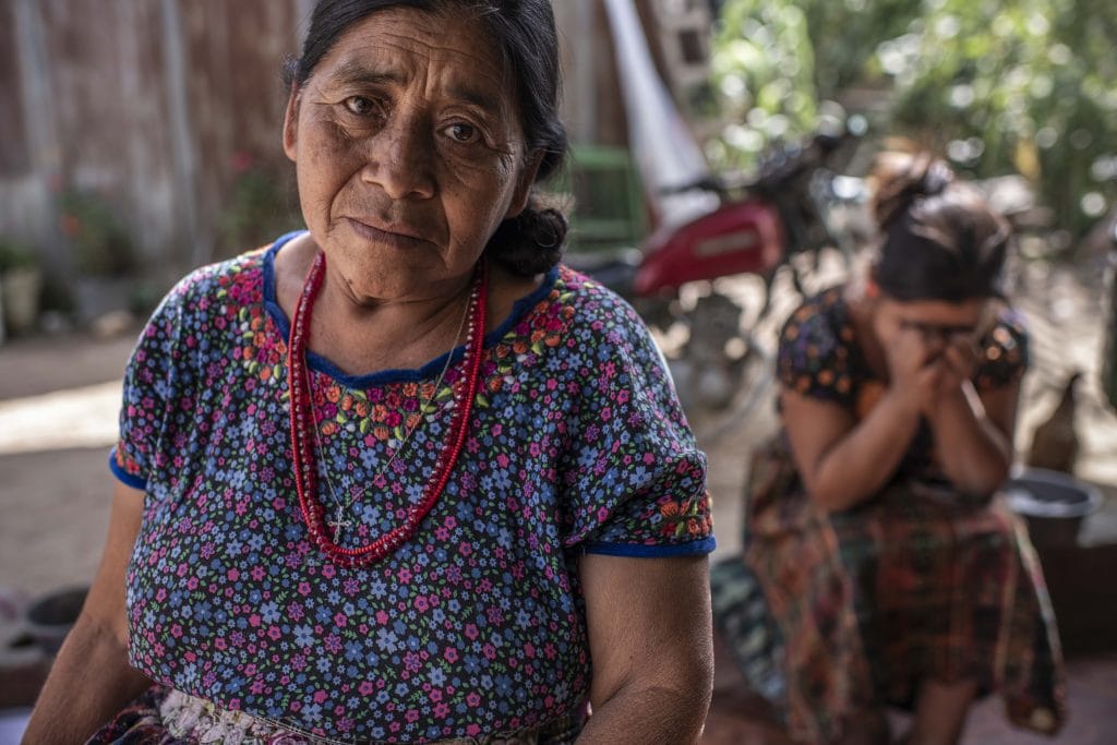 Older woman in a floral dress with a red beaded necklace sits outdoors, looking at the camera; a second woman in the background covers her face.