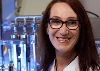 Smiling female doctor or scientist wearing a white lab coat and purple glasses, in a laboratory setting with lab equipment in the background (Texas Children's Hospital logo visible).