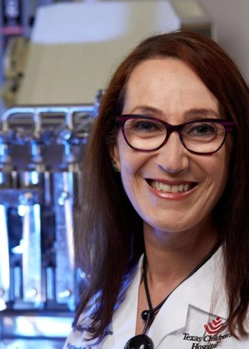 maria elena botazzi cientifica hondureña – SoyMigrante.com REVISTA Smiling female doctor or scientist wearing a white lab coat and purple glasses, in a laboratory setting with lab equipment in the background (Texas Children's Hospital logo visible).