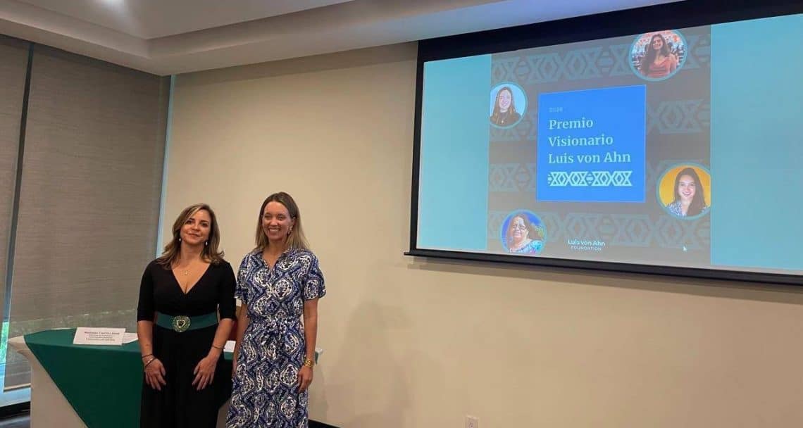 Two women pose for a photo in front of a projected slide announcing the Premio Visionario Luis von Ahn award in a conference room.