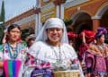 Central woman in white traditional dress smiling, holding a wicker basket during a cultural festival with colorful outfits in the background.