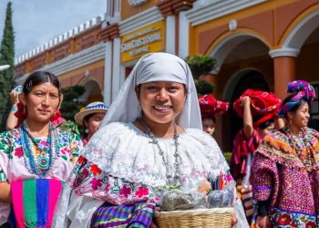 Central woman in white traditional dress smiling, holding a wicker basket during a cultural festival with colorful outfits in the background.
