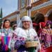 Central woman in white traditional dress smiling, holding a wicker basket during a cultural festival with colorful outfits in the background.