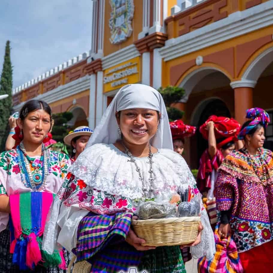 Central woman in white traditional dress smiling, holding a wicker basket during a cultural festival with colorful outfits in the background.