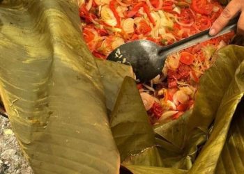 Person scooping bright chopped vegetables (onions, peppers, tomatoes) from a large banana leaf–wrapped dish with a spoonble