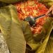 Person scooping bright chopped vegetables (onions, peppers, tomatoes) from a large banana leaf–wrapped dish with a spoonble