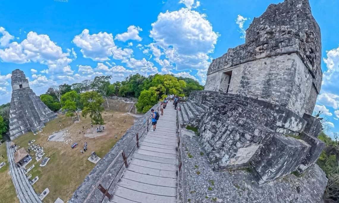 Tourists walk along a raised wooden walkway between ancient Mayan temple ruins under a bright blue sky.