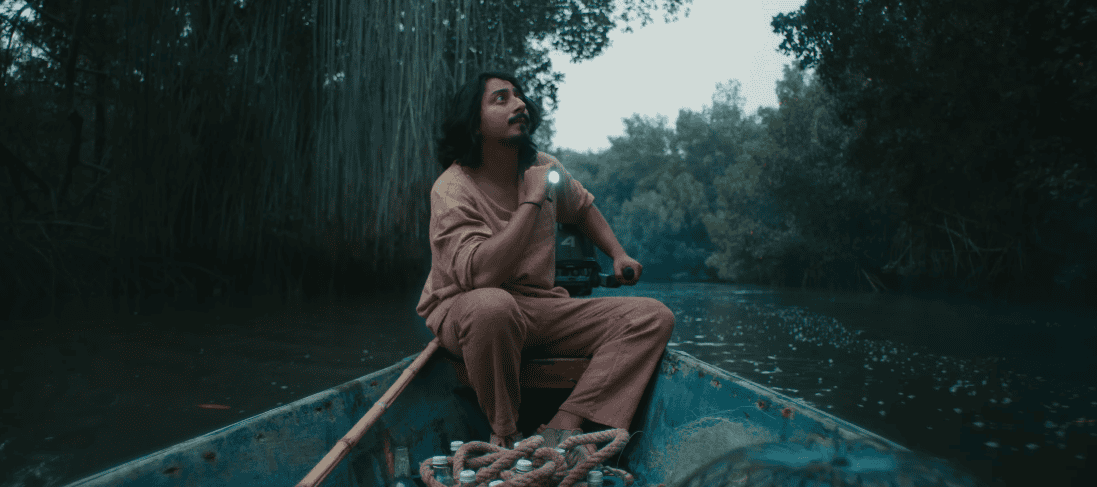 Man in a beige shirt and brown pants sits in a small boat on a river, holding a lit flashlight and looking upward toward the trees ahead.