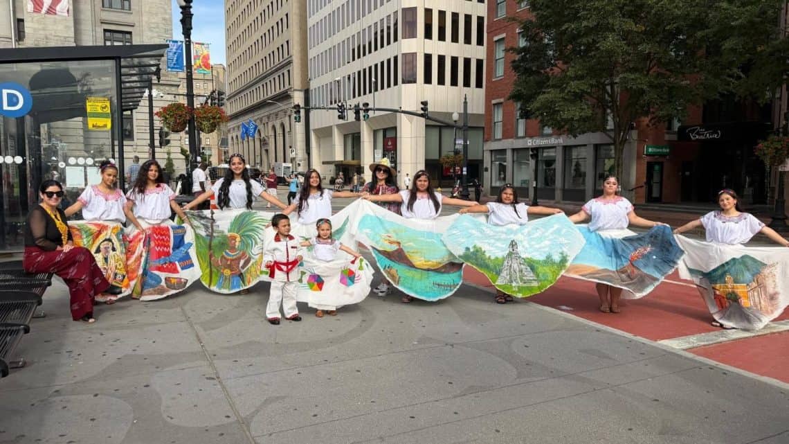 Group of people in white shirts holding a long illustrated cloth displaying colorful landscapes, posing on a city street during a parade or festival.