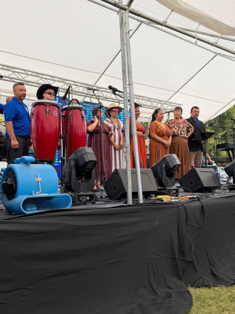 Diverse group of performers on an outdoor stage under a white canopy, singing and playing red conga drums with microphones and stage gear nearby.