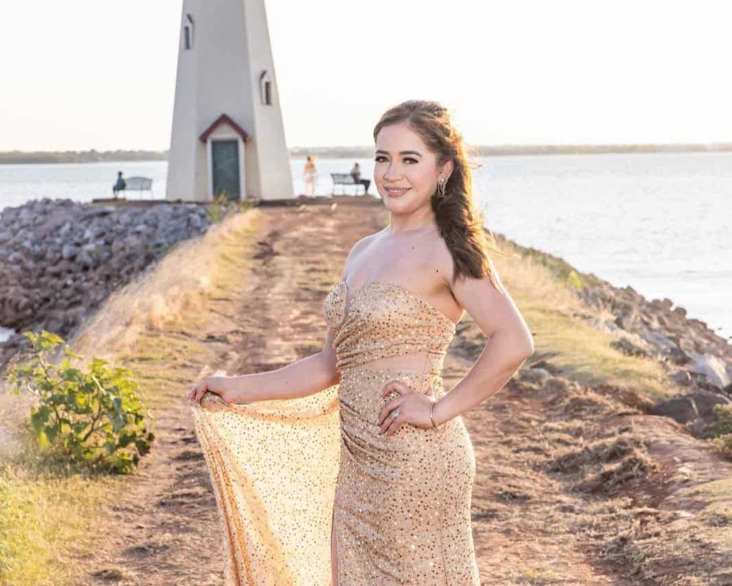 Woman in a gold sequined strapless gown posing on a rocky waterfront path with a lighthouse in the background.