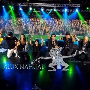 Group of performers on stage posing for a photo with a cheering crowd and colorful stage lights behind them.
