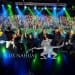 Group of performers on stage posing for a photo with a cheering crowd and colorful stage lights behind them.