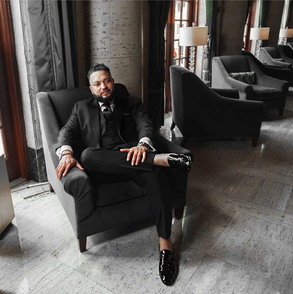 Man in a black suit relaxing on a dark chair in a luxurious lounge with other armchairs and lamps in the background.