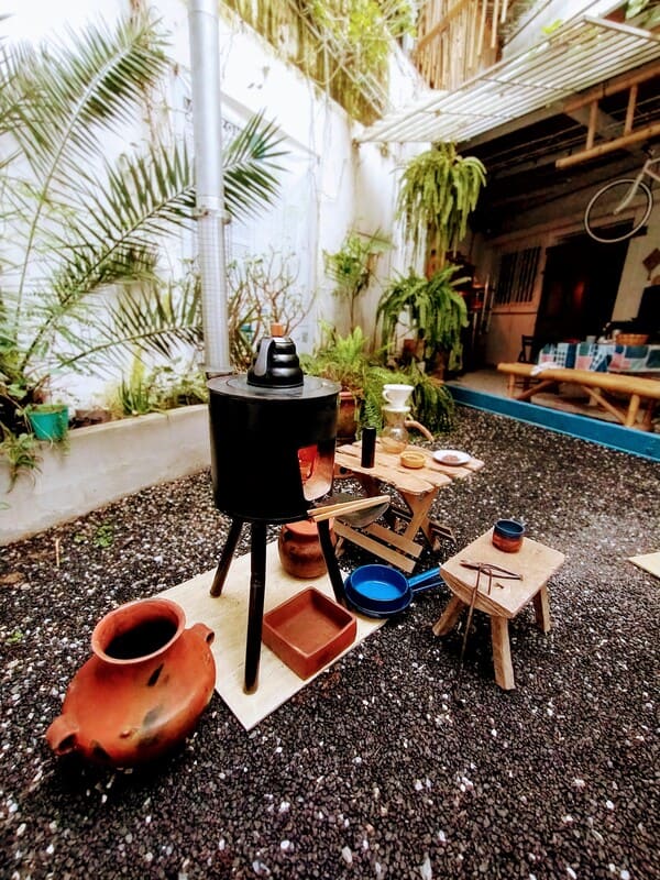 Outdoor courtyard scene with a black outdoor stove, clay pot, and small wooden tables set with bowls and cups among potted plants and gravel ground.
