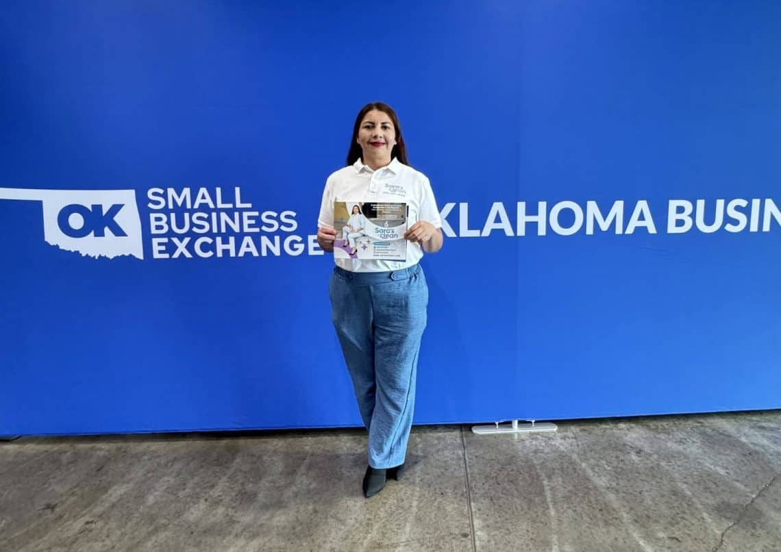 Woman standing in front of a blue Oklahoma Small Business Exchange banner, holding a flyer and smiling.