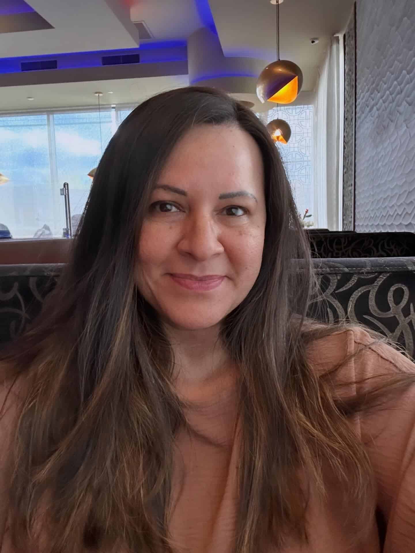 Close-up of a smiling woman with long dark hair in a casual indoor setting, pendant lights and patterned seating in the background.