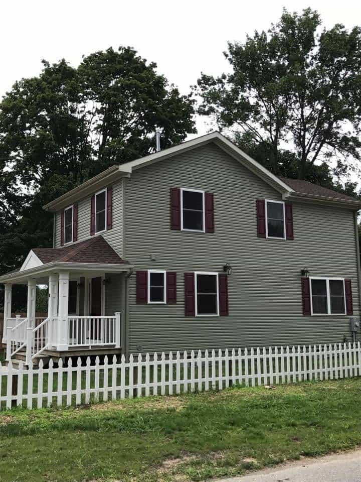 Two-story gray house with maroon shutters, white front porch, and a white picket fence in a grassy yard. Behind are tall trees.