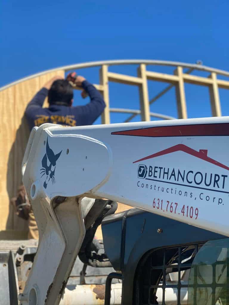 Construction worker in a blue hoodie framing a wooden wall on a sunny construction site, with a Bethancourt Construction boom lift in the foreground.