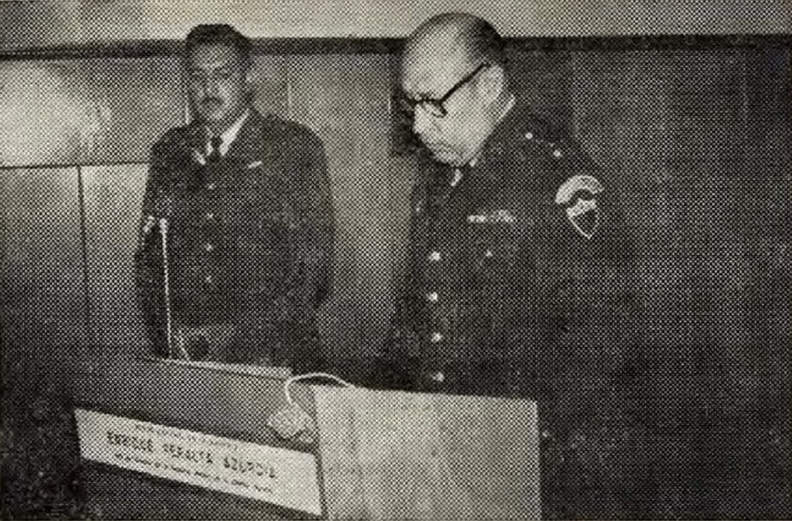 Discurso del coronel Enrique Peralta Azurdia Jefe de Gobierno acerca de las actividades del Ejército en 1965 año en que se aprobó la nueva Constitución que estaría vigente hasta 1982 – SoyMigrante.com REVISTA – SoyMigrante.com REVISTA Two uniformed police officers stand at a podium in a room, one looking down at papers.