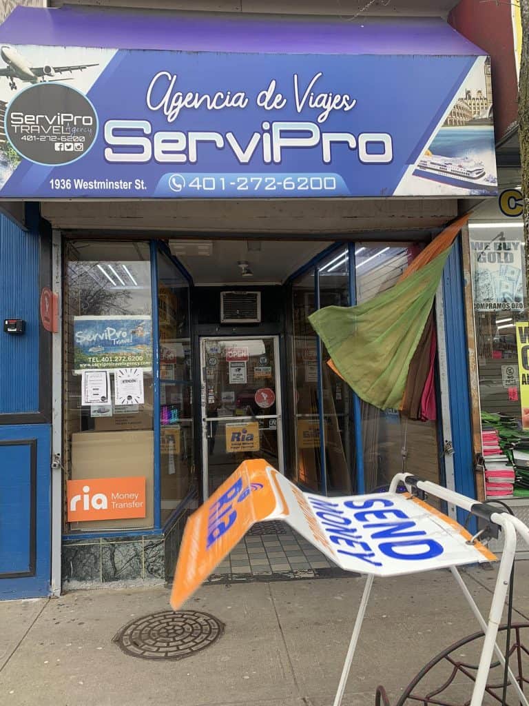 Front of a travel agency storefront with a large blue sign saying 'Agencia de Viajes ServiPro' over a glass entrance and posters in the windows; colorful fabric hangings to the right side of the doorway.