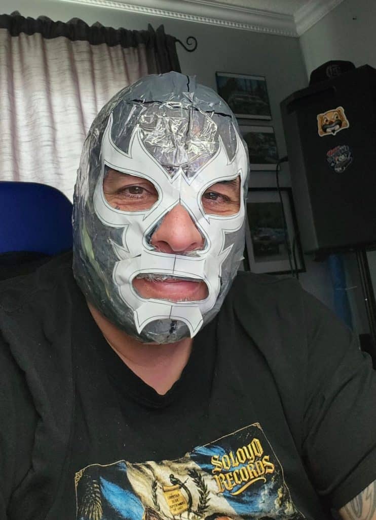 Person wearing a shiny silver Lucha Libre mask indoors, facing the camera with a black shirt featuring a Slow Records graphic