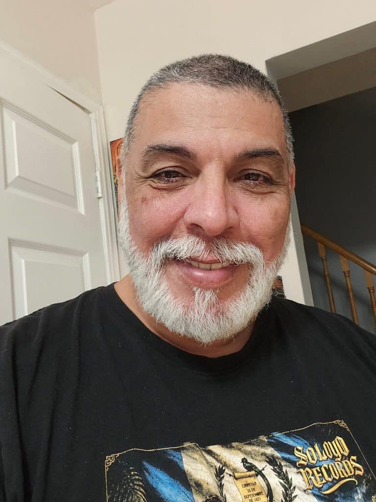 Close-up selfie of a smiling man with gray beard and short gray hair, wearing a black t-shirt indoors near a doorway and staircase.