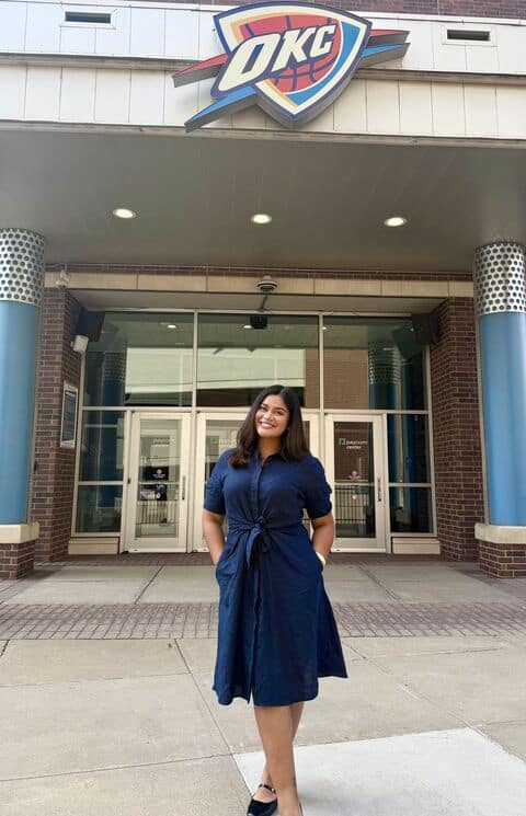 Gabriela Ulloa se unió recientemente al área de comunicación y relaciones públicas del equipo de la NBA, Oklahoma City Thunder. – SoyMigrante.com REVISTA – SoyMigrante.com REVISTA Woman in a navy blue dress posing outside the OKC entrance, smiling at the camera.