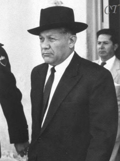 Black-and-white photo showing a middle-aged man in a dark suit and fedora walking, with a uniformed officer nearby and a serious expression.