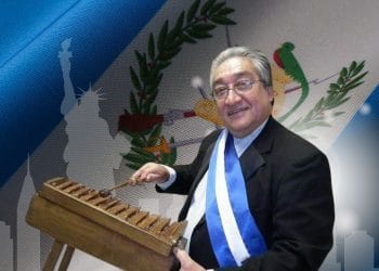 Older man in a suit wearing a blue-and-white ceremonial sash plays a wooden xylophone on a decorated backdrop.
