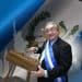 Older man in a suit wearing a blue-and-white ceremonial sash plays a wooden xylophone on a decorated backdrop.